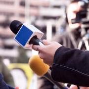 A reporter holds her microphone up to a source as a cameraman takes in the scene in the background.