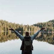 Girl wearing a beanie holding her arms up with joy in front of a forested lake.