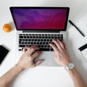 a person working on a laptop with a notebook, pen, phone and an orange nearby