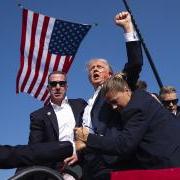 Donald Trump with an extended arm and agents surrounding him, stands on a stage, American flag draped from above, stands with streams of blood across his face.