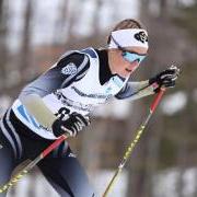 Nordic skier Hanna Abrahamsson wears a CU headband, sunglasses and a competition jersey, in a racing position with poles in hand, and small tree trunks in the background.