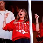 Taylor Swift cheering in a stadium box wearing a Kansas City Chiefs sweatshirt